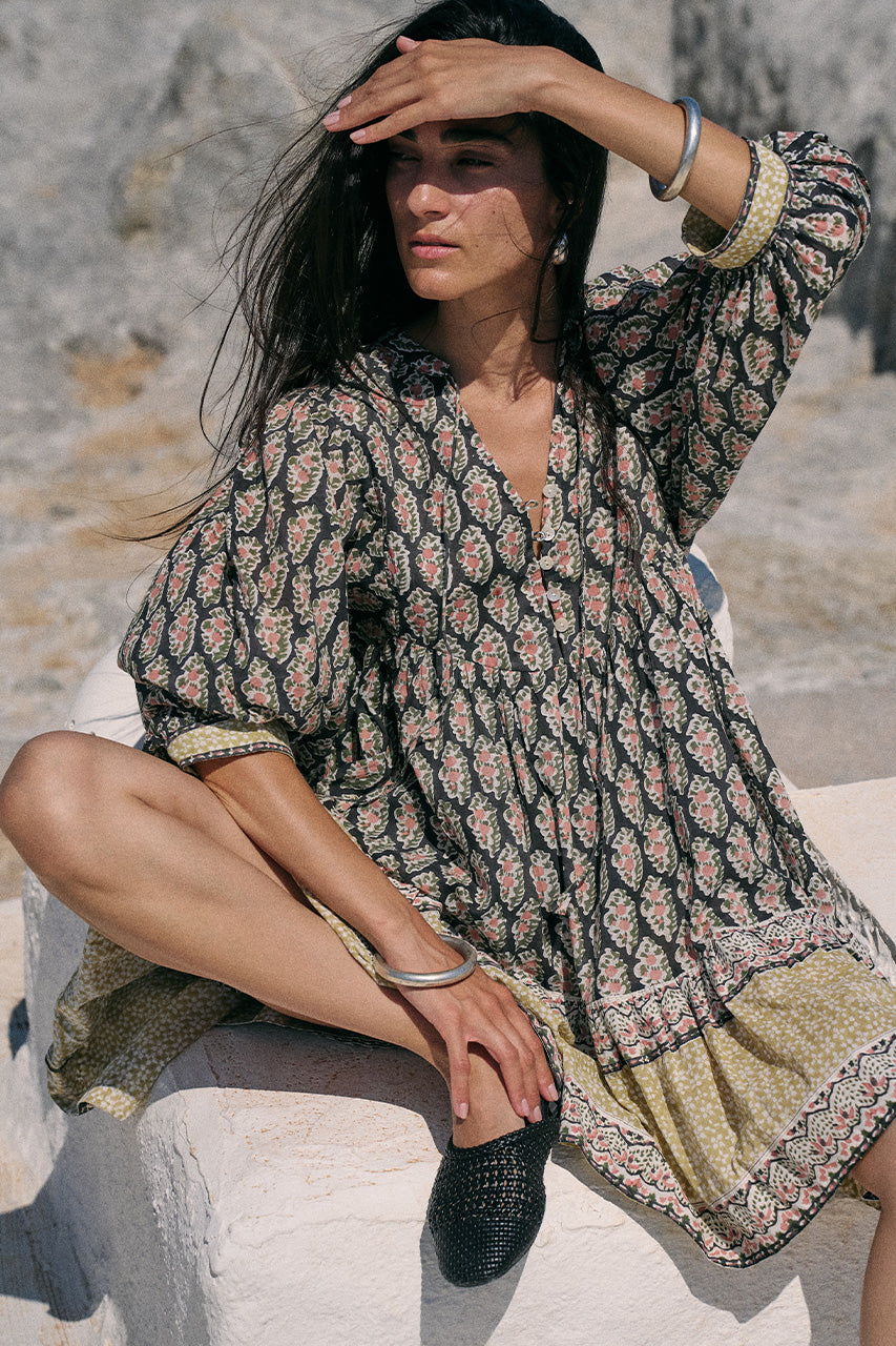 Woman seated on white stone wearing the Daughters of India Bhoomi Mini Dress in Onyx, shielding her eyes from the sun, showcasing the paisley block print cotton fabric