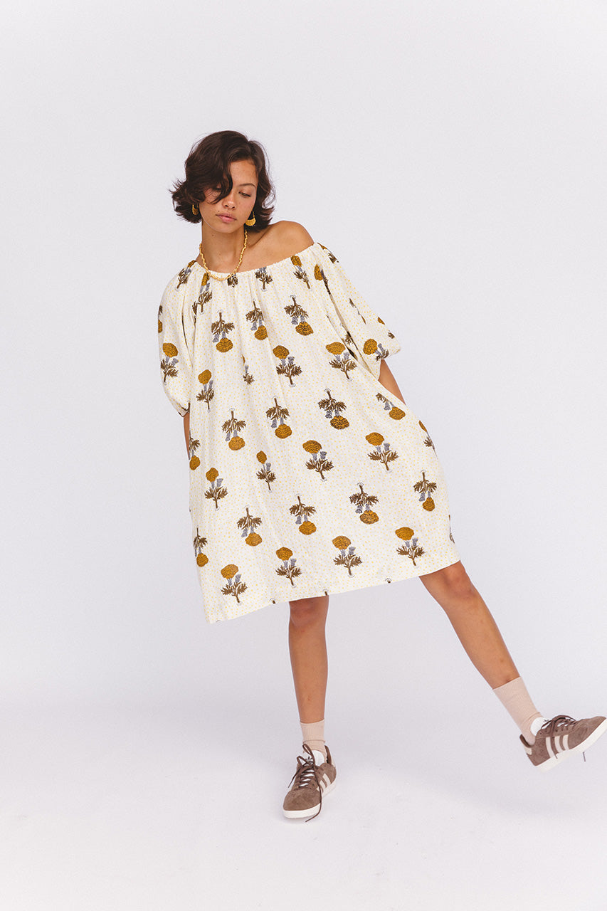 Dahlia Mini Dress Marigold — playful studio shot, leg kicked to side, off-shoulder, showing A-line linen mini silhouette, white with golden amber marigold hand block print, ethical slow fashion | Daughters of India