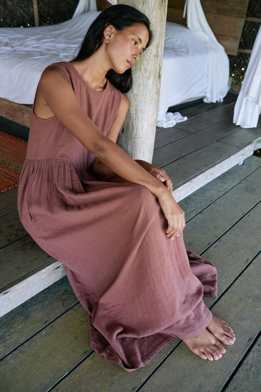 Nadi Dress Chocolate — seated on wooden floor leaning against post, chocolate brown double gauze cotton midi dress flowing over legs, rustic bamboo bedroom with white bedding | Daughters of India