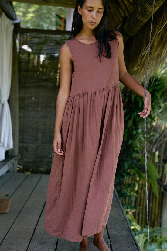 Nadi Dress Chocolate — full length front view on wooden deck under thatched roof, chocolate brown double gauze cotton midi dress with round neckline and gathered waist, tropical garden | Daughters of India